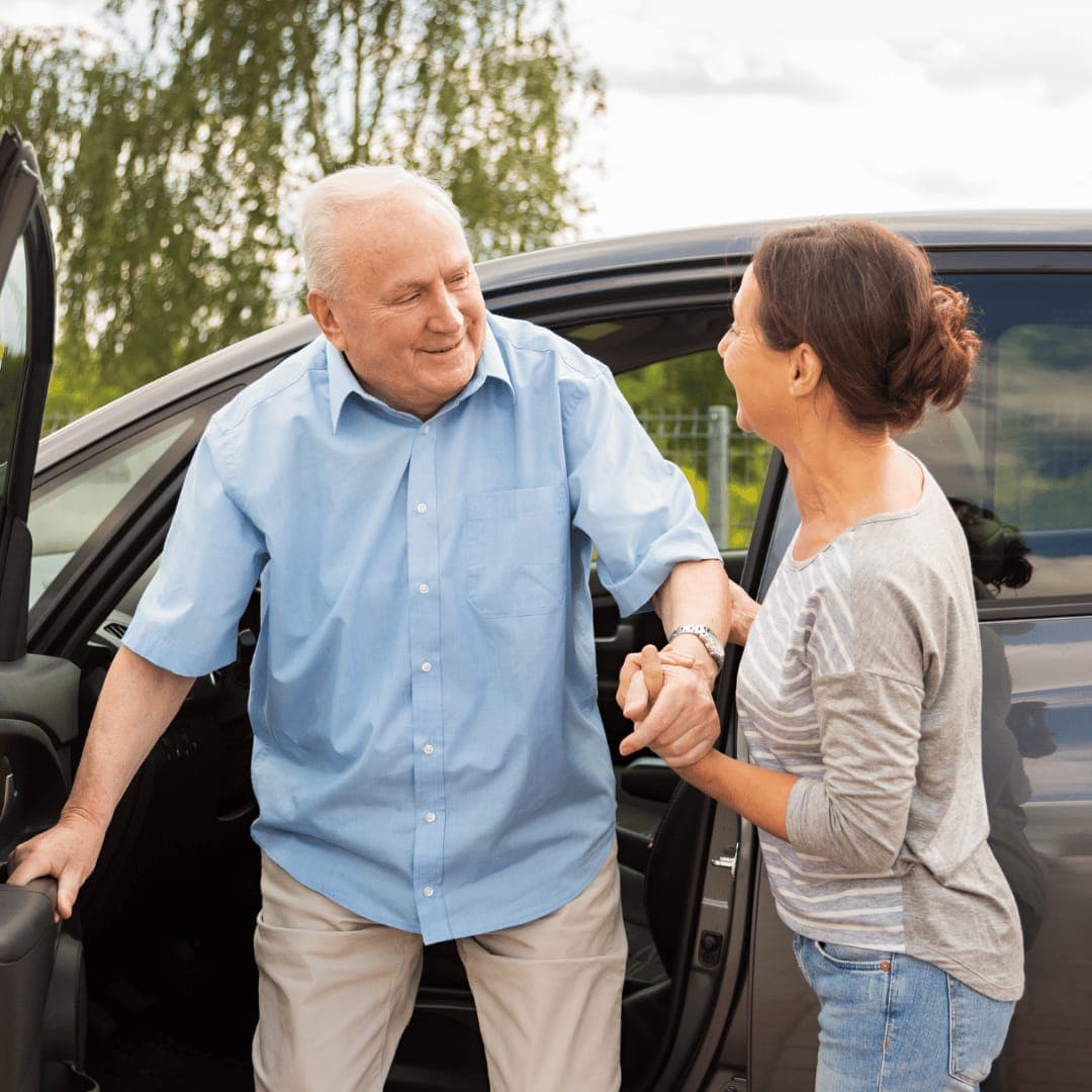A helper supporting an older man as he steps out of a car